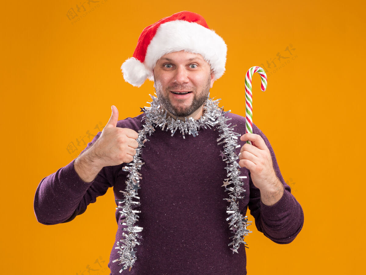 拇指令人印象深刻的中年男子戴着圣诞帽 脖子上戴着金箔花环 手里拿着圣诞甜手杖 看着相机 在橙色背景上孤立地竖起大拇指圣诞节手杖帽子