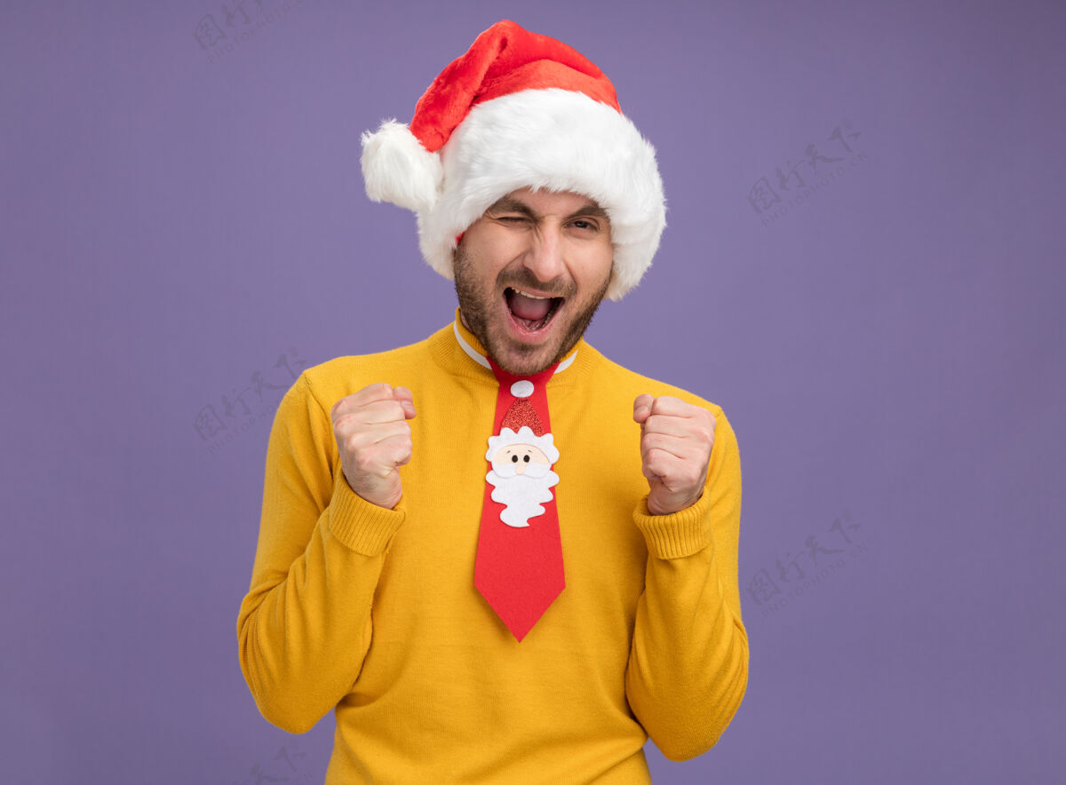 年轻快乐的年轻白种人戴着圣诞帽打着领带眨眼做着“是”的手势隔离在紫色的墙上 留着复制空间领带手势帽子