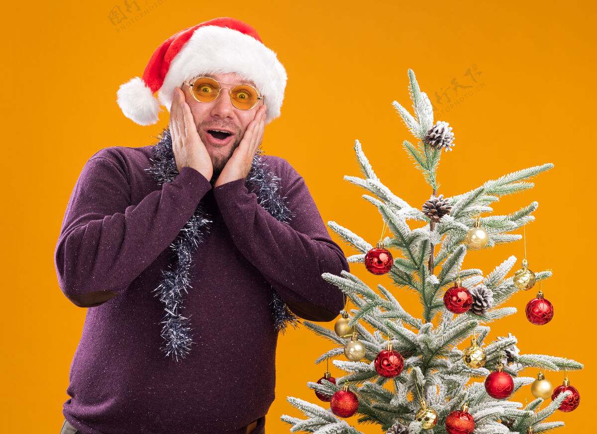 保持兴奋的中年男子戴着圣诞帽 脖子上戴着金箔花环 戴着眼镜 站在装饰好的圣诞树旁 手放在脸上 看着橙色背景上的相机帽子周围眼镜
