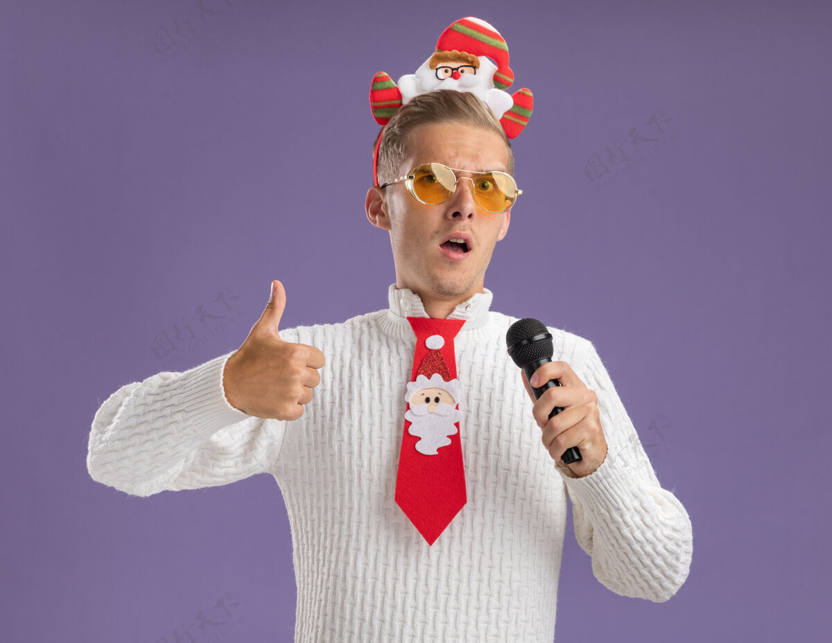 圣诞快乐自信的年轻帅哥戴着圣诞老人的头带和领带戴着眼镜拿着麦克风看着摄像机在紫色背景上孤立地竖起大拇指相机拇指快乐