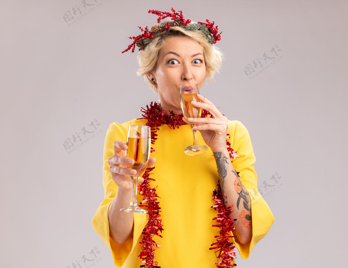 花环年轻的金发女郎戴着圣诞花环 脖子上戴着金箔花环 手里拿着两杯香槟 喝着一杯 隔离在白色墙上 留着复印空间喝酒花环头