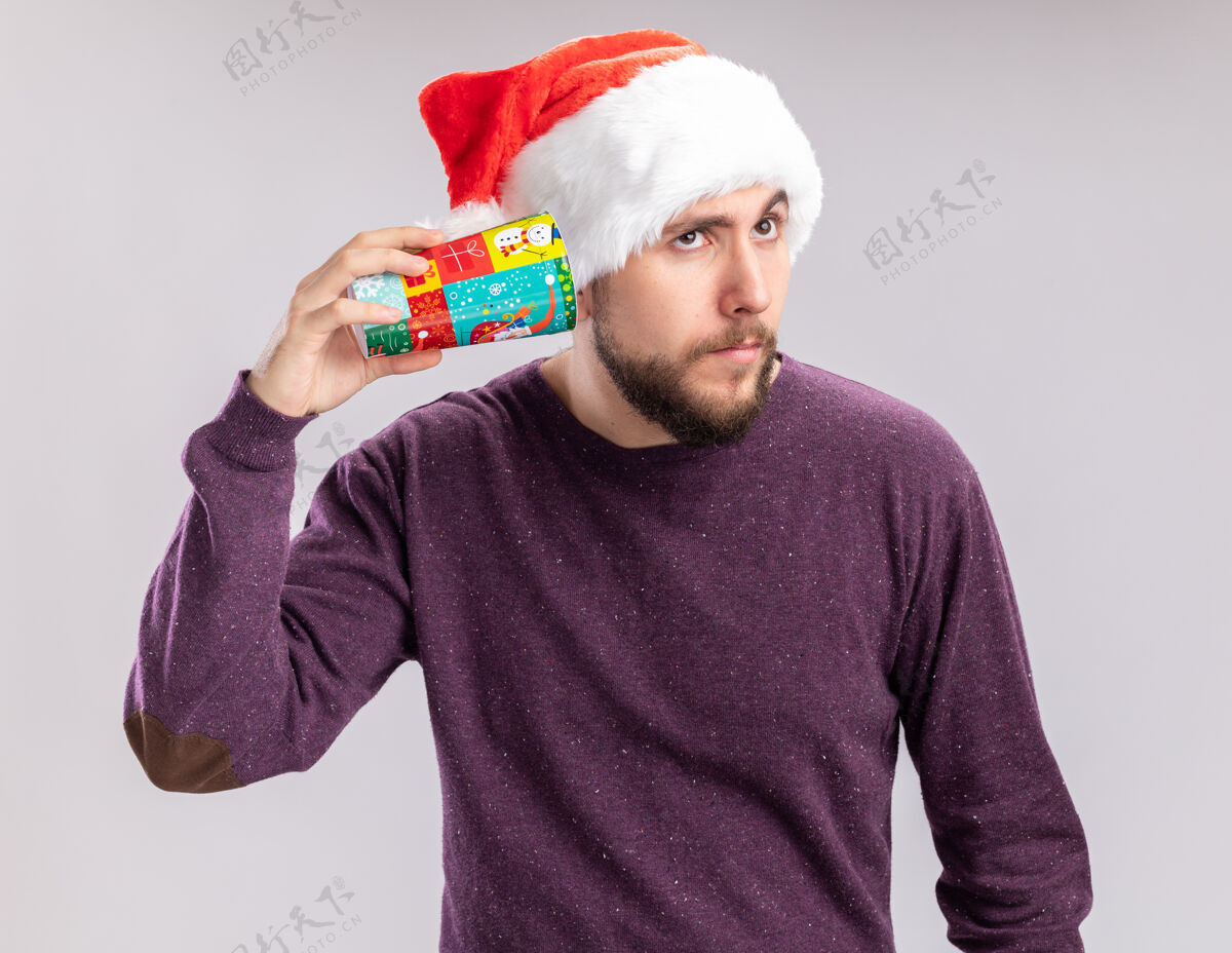 圣诞老人一个穿着紫色毛衣 戴着圣诞帽的年轻人把五颜六色的纸杯捂在耳朵上 想听听站在白色背景上的声音年轻人毛衣帽子