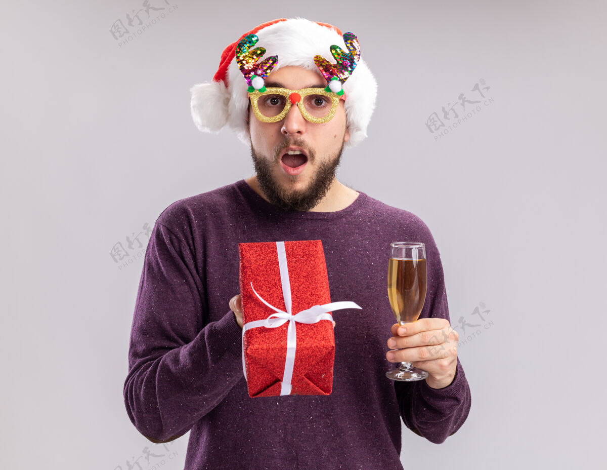 年轻年轻人穿着紫色毛衣 戴着圣诞帽 戴着搞笑的眼镜 手里拿着一杯香槟 惊讶地看着镜头 新年假期的概念站在白色的背景上年站着圣诞老人