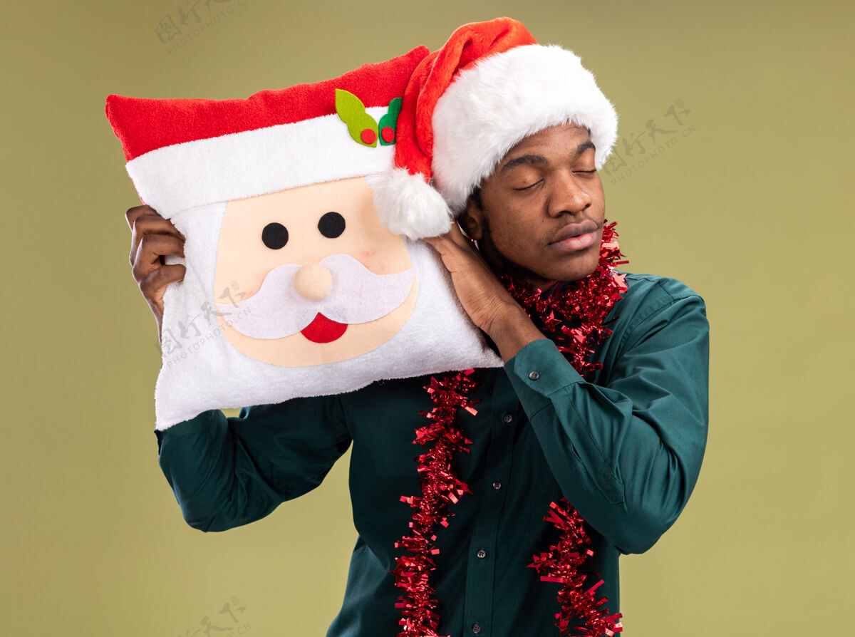圣诞节戴着圣诞帽 戴着花环 抱着圣诞枕头 闭着眼睛站在绿色背景上的非裔美国人枕头举行眼睛