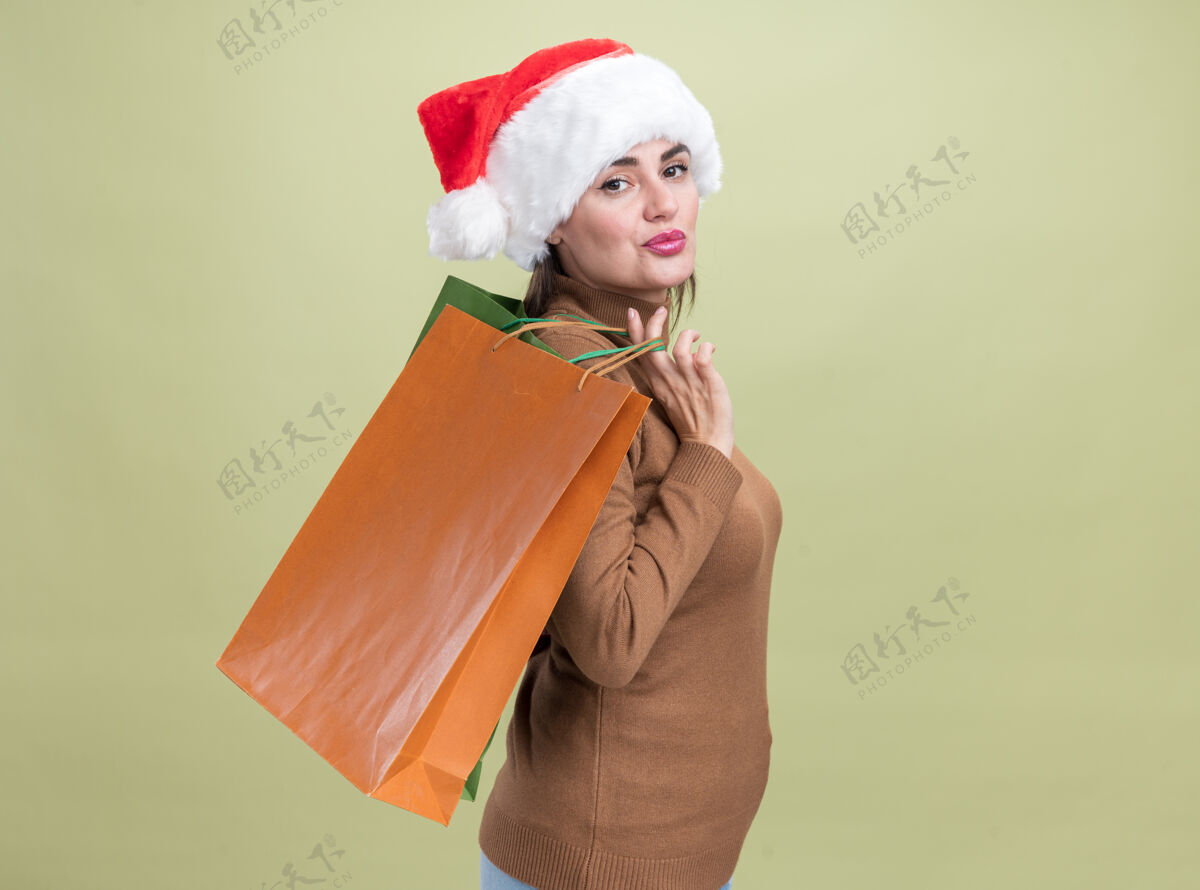 年轻高兴地站在侧视图年轻漂亮的女孩戴着圣诞帽手持礼品袋在肩上孤立的橄榄绿背景查看圣诞节包