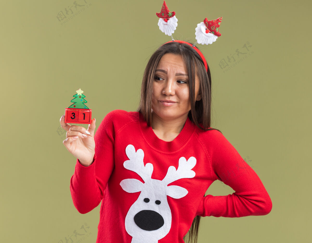 圣诞节印象深刻的年轻亚洲女孩戴着圣诞发箍举行 并在圣诞节玩具看把手放在臀部隔离在橄榄绿的背景推杆臀部年轻