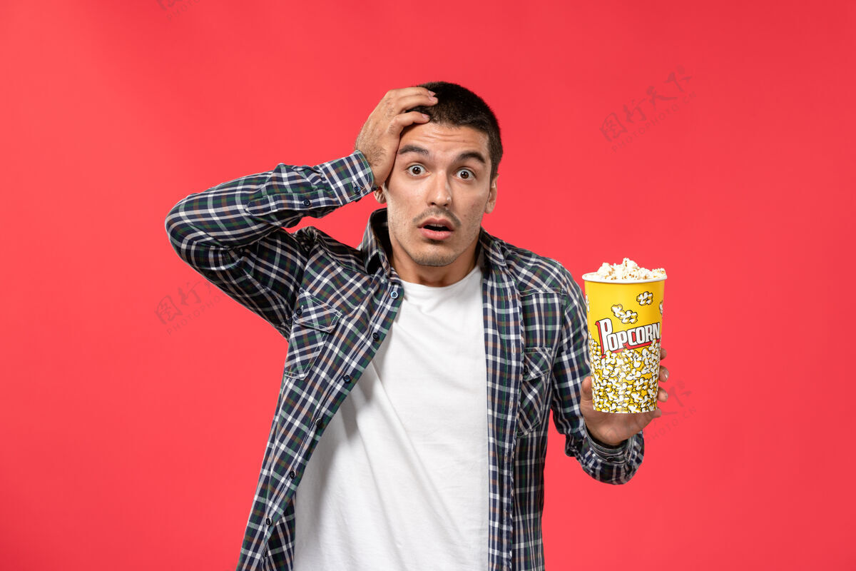 观点正面图年轻男子拿着爆米花包 表情惊讶地站在淡红色的墙上看男性电影电影年轻包装