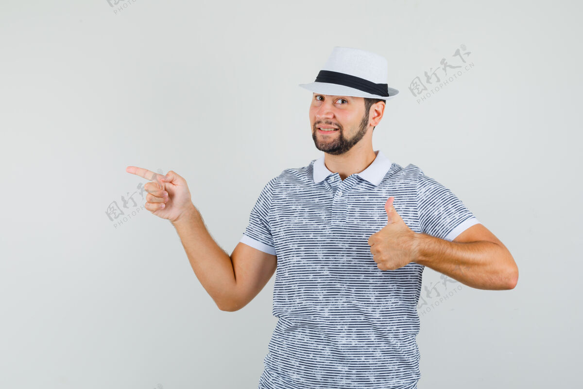 衬衫年轻人指着别处 同时显示拇指在条纹t恤衫 帽子和看起来高兴 前视图男人休闲时尚