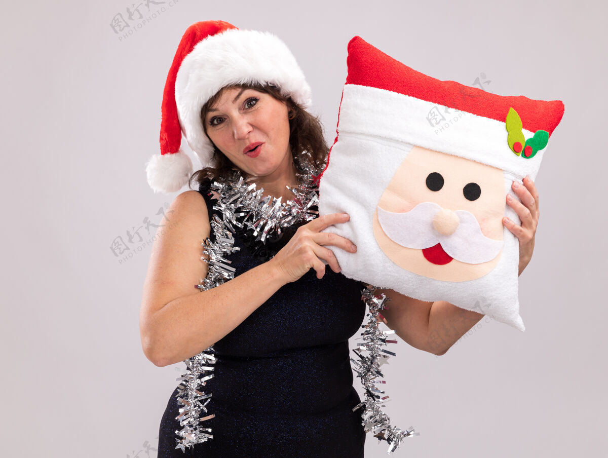 印象深刻印象深刻的中年妇女戴着圣诞帽 脖子上戴着金箔花环 抱着圣诞老人的枕头 看着隔离在白色背景上的相机圣诞老人抱抱枕头