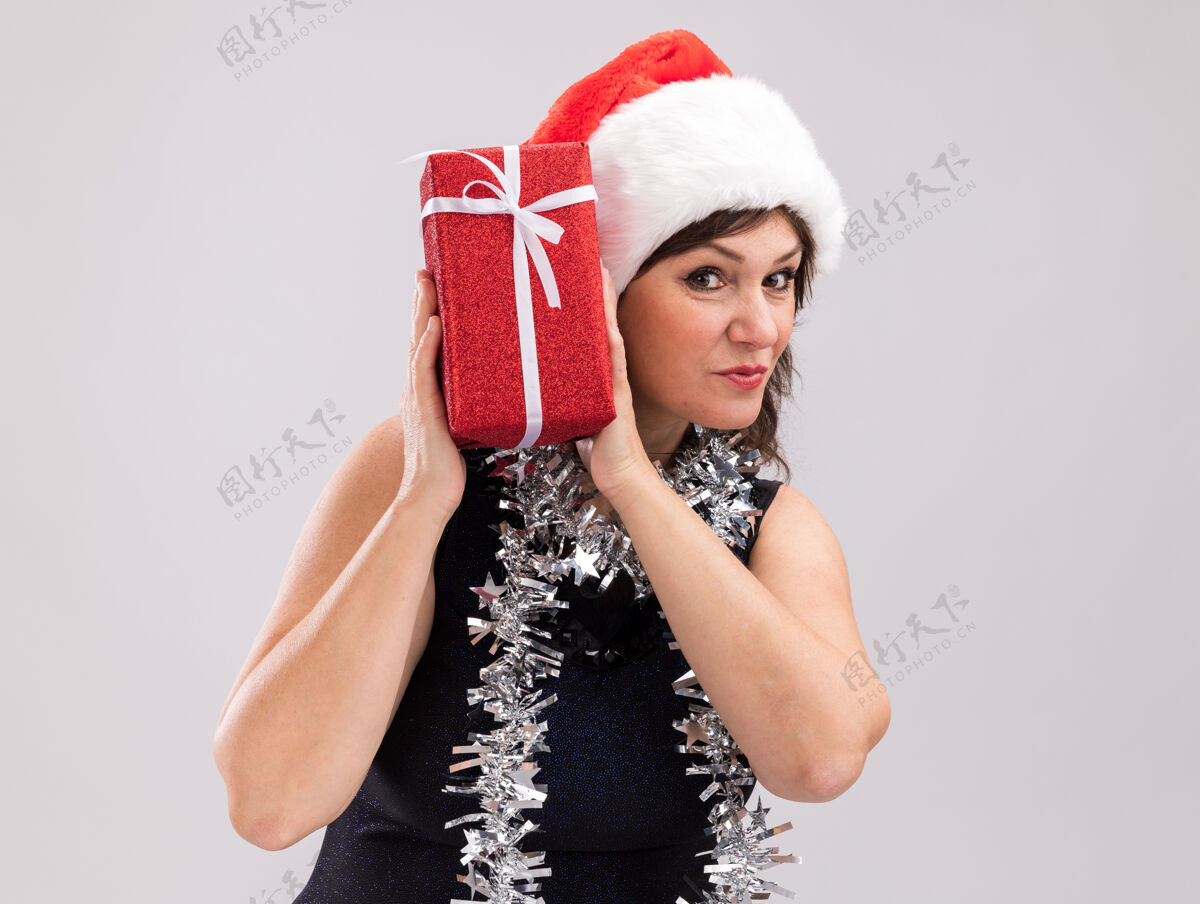 周围好奇的中年妇女戴着圣诞帽 脖子上戴着金箔花环 头上拿着圣诞礼包 看着隔离在白色背景上的相机 还有复印空间圣诞节金属丝帽子
