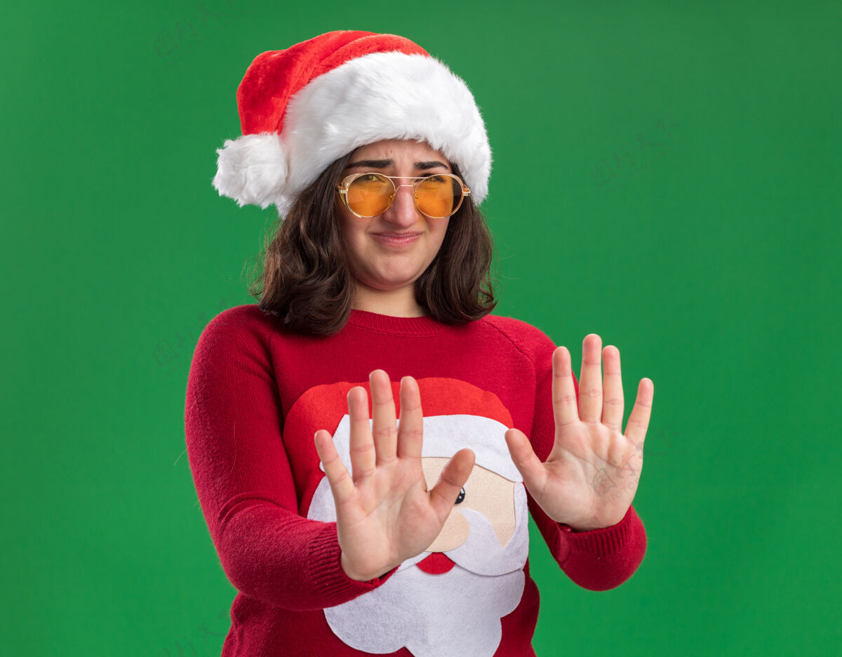 女孩穿着圣诞毛衣 戴着圣诞帽 戴着眼镜的年轻女孩站在绿色的背景下 厌恶地看着镜头 手伸出毛衣站着圣诞节