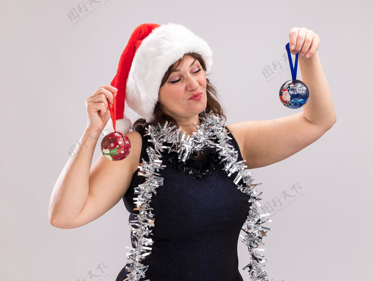 帽子可疑的中年妇女戴着圣诞帽 脖子上戴着金属丝花环 手里拿着圣诞饰品 看着其中一件孤立在白色背景上的圣诞饰品花环可疑金属丝