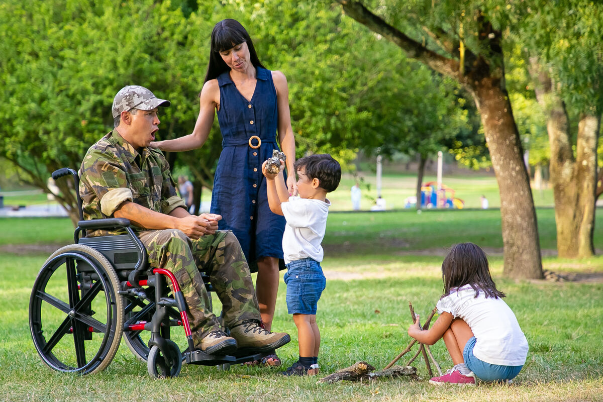 侧视图孩子们在母亲和残疾军人父亲坐在轮椅旁的公园里为营火布置木柴男孩向兴奋的父亲展示木柴残疾退伍军人或家庭户外概念男人视图完整