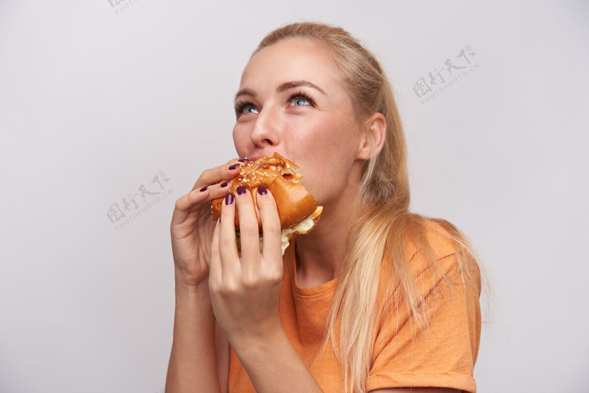 食物年轻漂亮的蓝眼睛金发女郎的特写镜头 随意的发型 手里拿着美味的汉堡 贪婪地咬着一块 站在白色的背景下室内休闲情绪