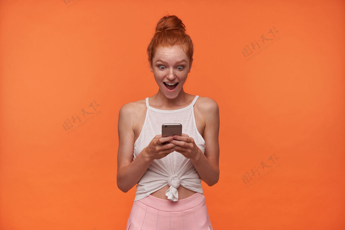 智能手机正面的年轻女读者头像 头发打结 手里拿着手机 惊奇地看着屏幕 扬起眉毛 张大嘴巴 孤立在橙色背景上20多岁抱着裙子
