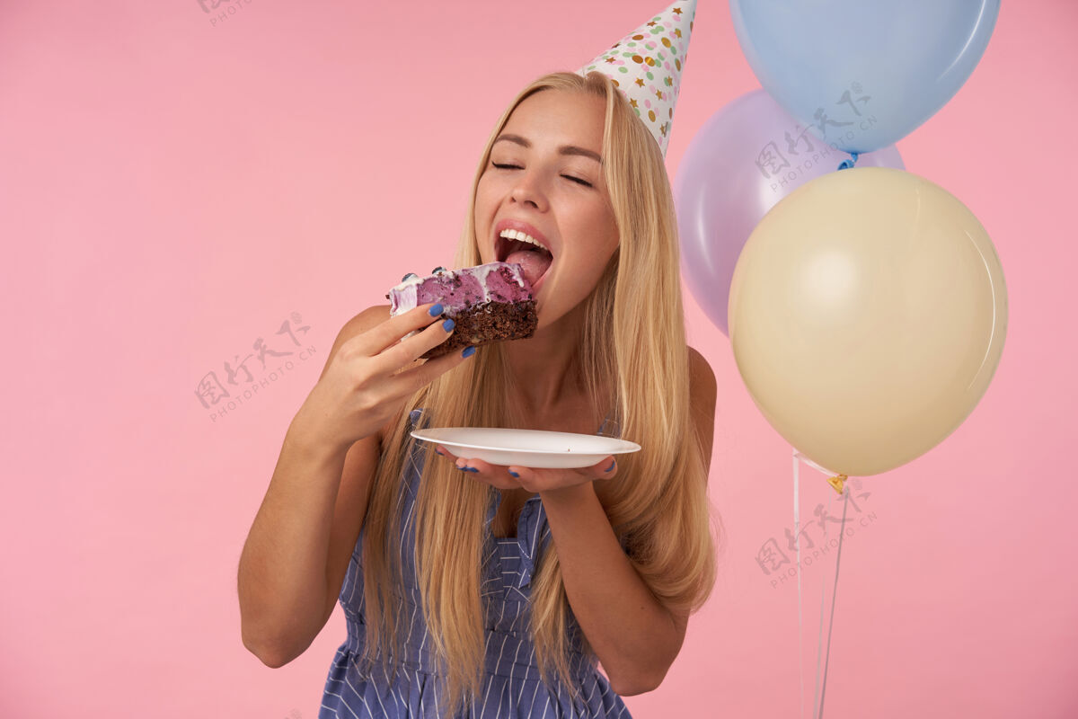 件身着节日服装的金发女郎手持一大块美味的蛋糕准备吃 手持五颜六色的气球庆祝生日 在粉色背景上摆姿势女士正面圆锥形