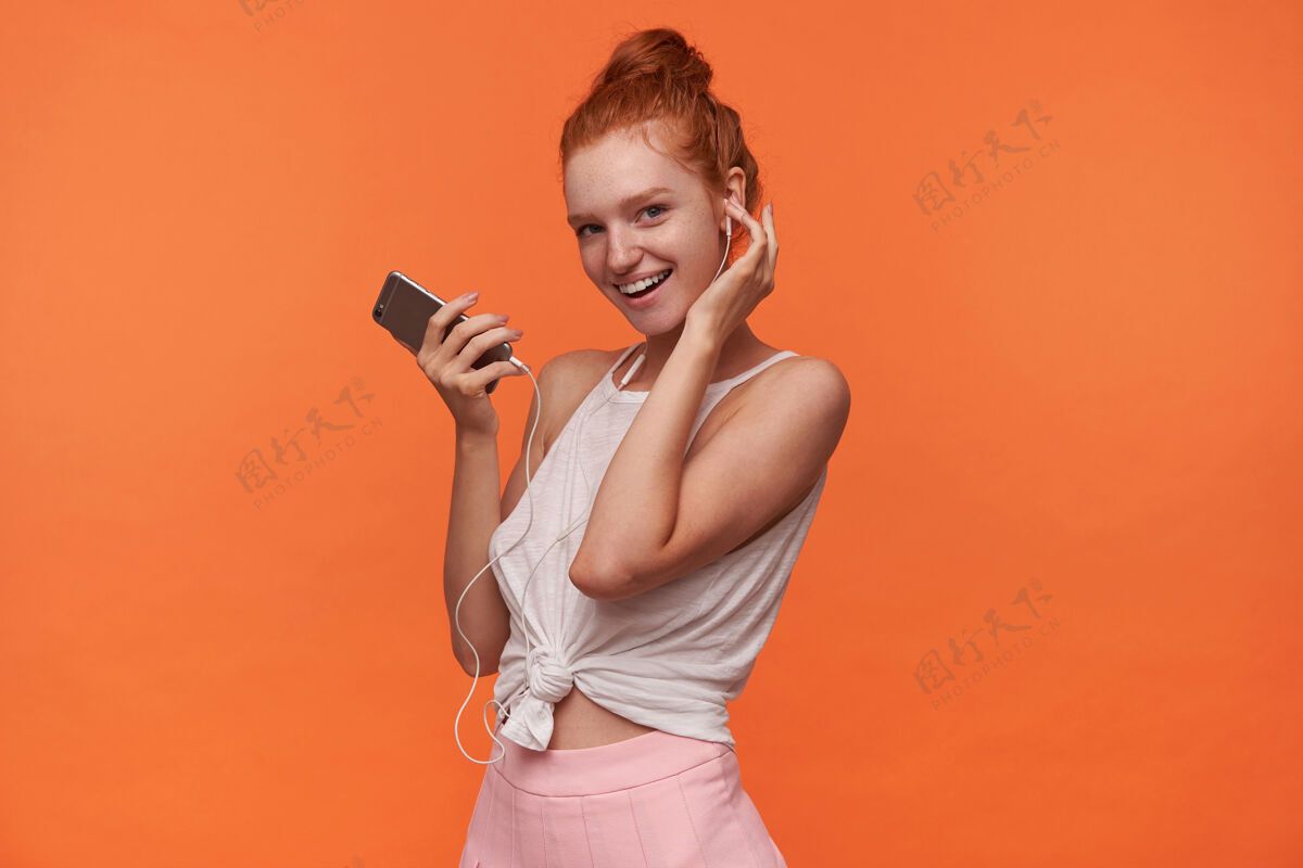 女士水平拍摄的是一位年轻漂亮的readhead女性 头发打结 穿着休闲服 面带微笑地看着相机 手里拿着智能手机 戴着耳机听音乐结发型手机