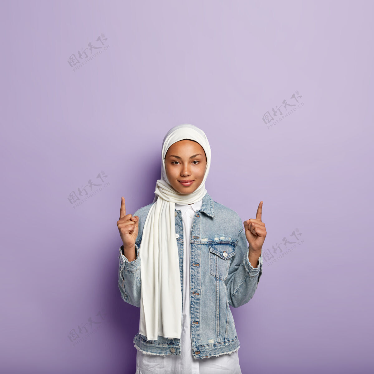 夹克深色皮肤的自信穆斯林妇女的垂直镜头 指向顶部 为顾客展示了巨大的向上复制空间 戴着白色丝巾和牛仔外套 隔离在紫色的墙上客户东方头巾