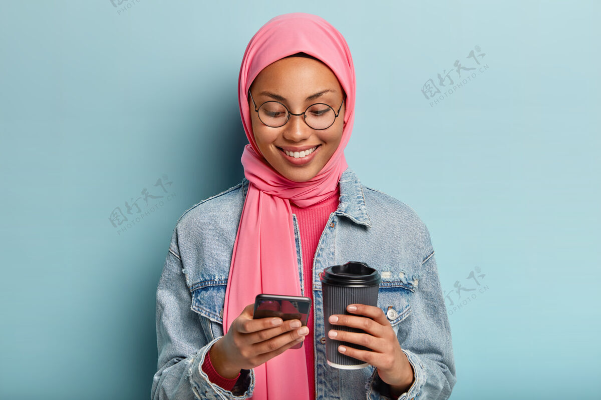 头巾快乐的混血女人 健康的深色皮肤 愉快的微笑 拿着手机和外卖咖啡 戴着粉色头巾 牛仔夹克 乐于接受信息 隔着蓝色的墙智能手机高兴微笑