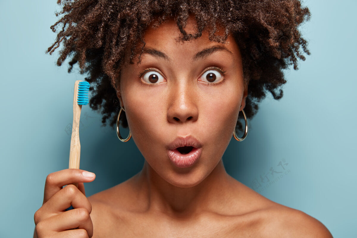 非洲惊讶的黑人妇女的特写肖像用窃听的眼睛盯着 张开嘴 拿着木刷 准备刷牙 惊讶于牙膏的效果 拥有健康的深色皮肤凝视新鲜年轻