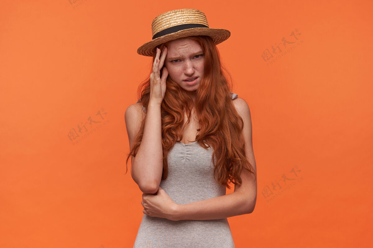 女士摄影棚拍摄的美丽的年轻红发女性 卷曲的狐狸精的头发藏在她的手掌后面 看着愧疚的脸 在橙色背景下孤立的相机休闲稻草长