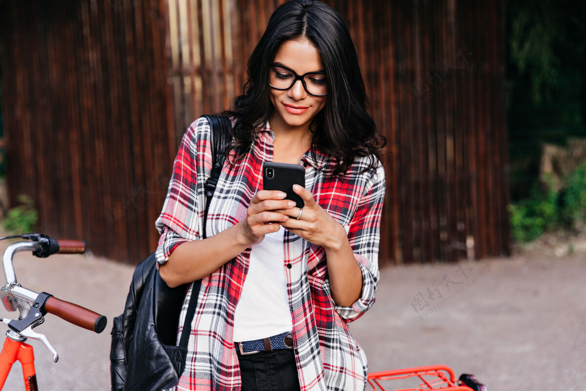 女人令人惊叹的拉丁女模特在等待某人时看着手机屏幕站在红色自行车旁的可爱黑发女人的户外肖像设备表情休闲