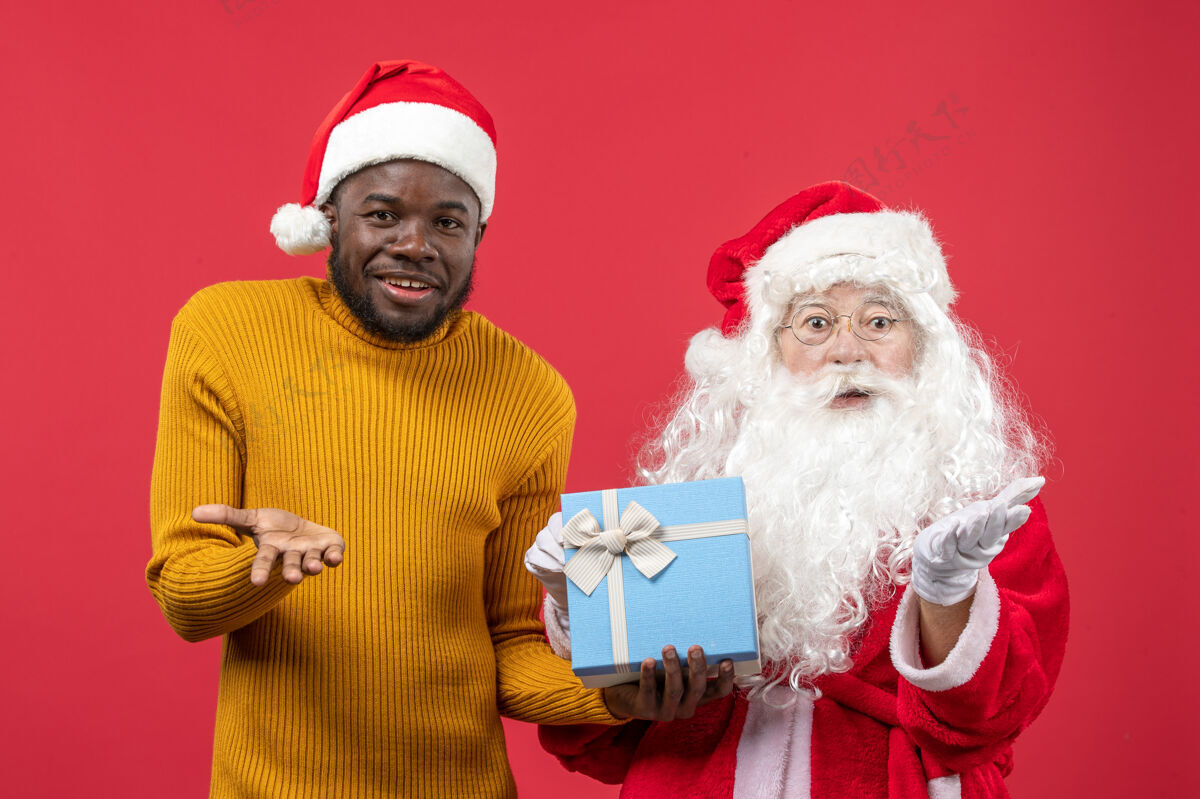 年轻红墙上的圣诞老人和拿着礼物的年轻人的正视图庆祝帽子礼物
