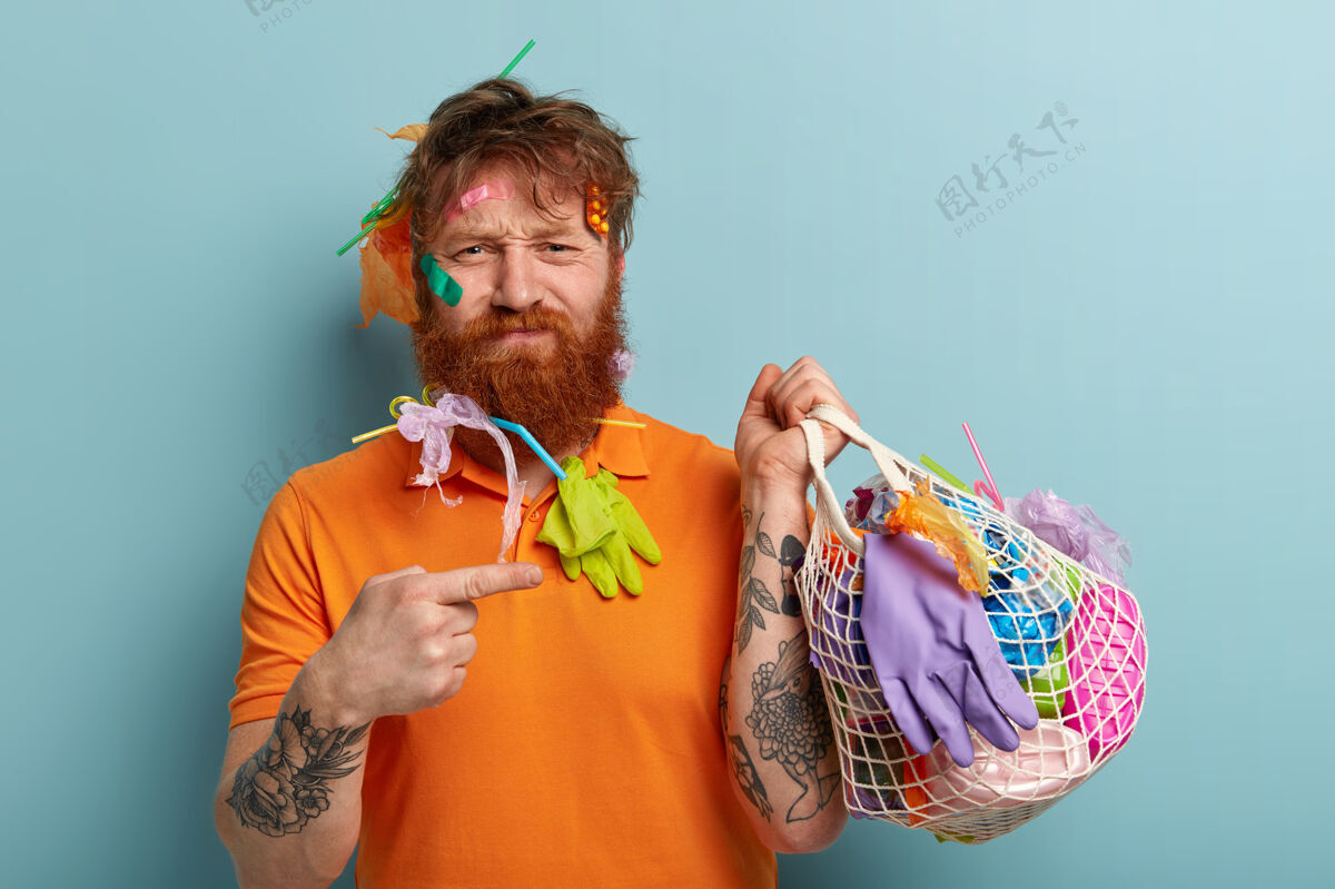 包装不满意的红发男人的照片 浓密的鬃毛 用食指指着装满塑料垃圾的袋子 穿着休闲的橙色t恤 手臂上有纹身 站在蓝色的墙上地球日垃圾垃圾生态