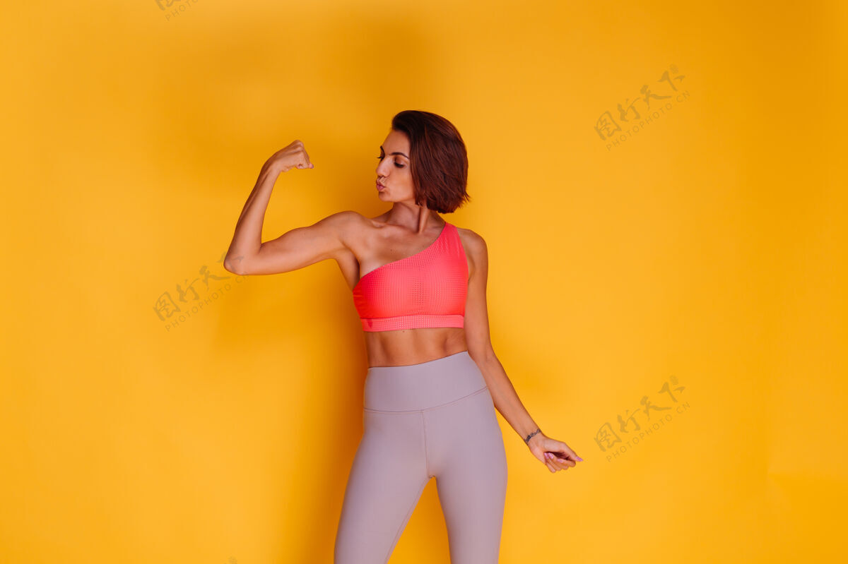 腹部年轻健壮的女人穿着运动服 时髦的上衣和紧身裤 对着黄色的墙壁摆姿势动机强壮活动