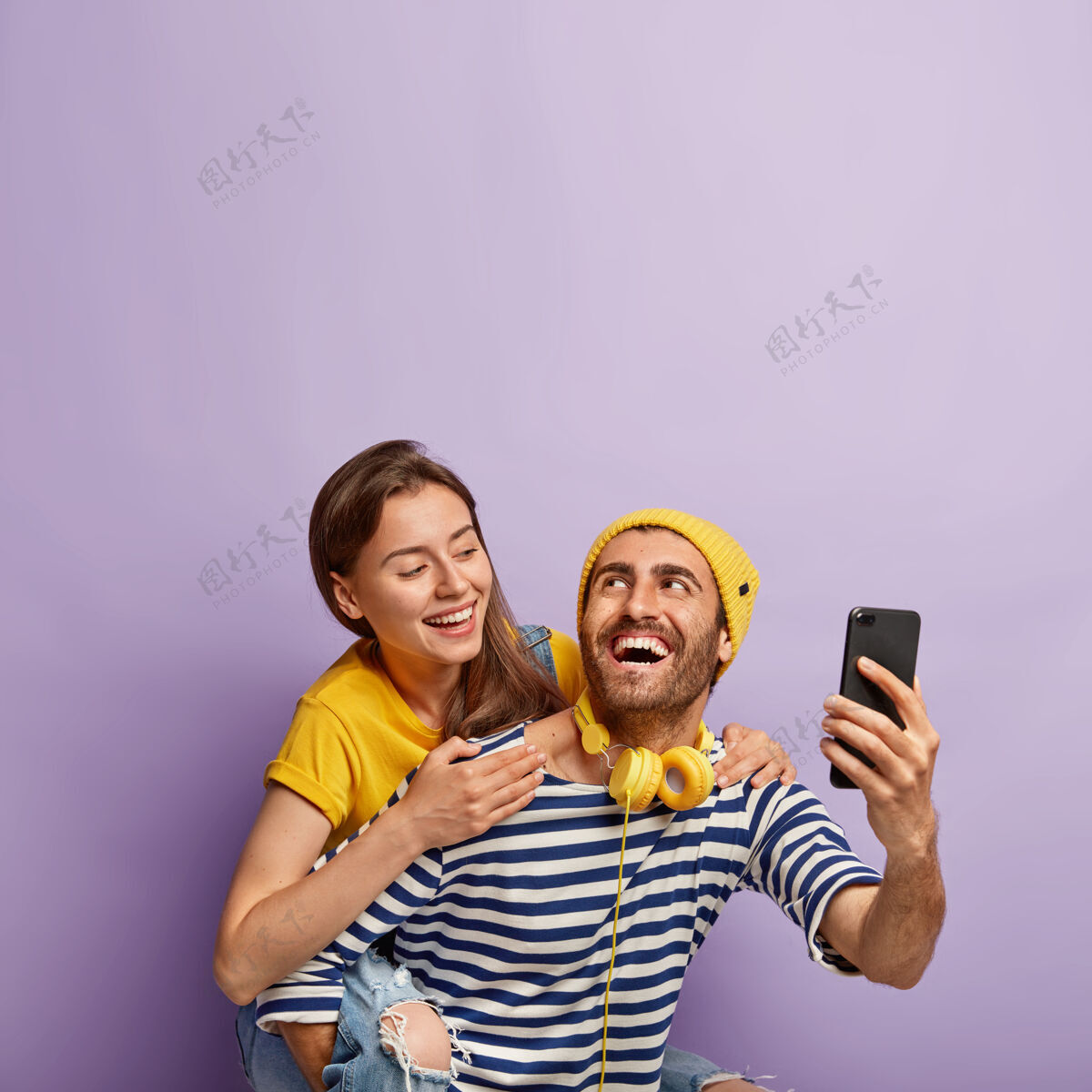 男朋友有趣的年轻夫妇在智能手机上自拍 享受骑在背上的乐趣 有快乐的表情 可爱的女人从背后拥抱男友 在紫色背景下孤立人骑拥抱摄影