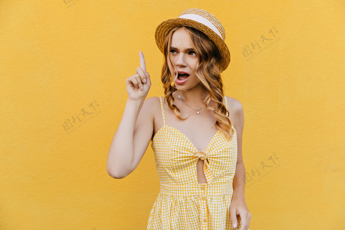 卷发有趣的女孩穿着时髦的黄色连衣裙摆姿势有兴趣的女模特戴着草帽环顾四周苗条黄色休闲