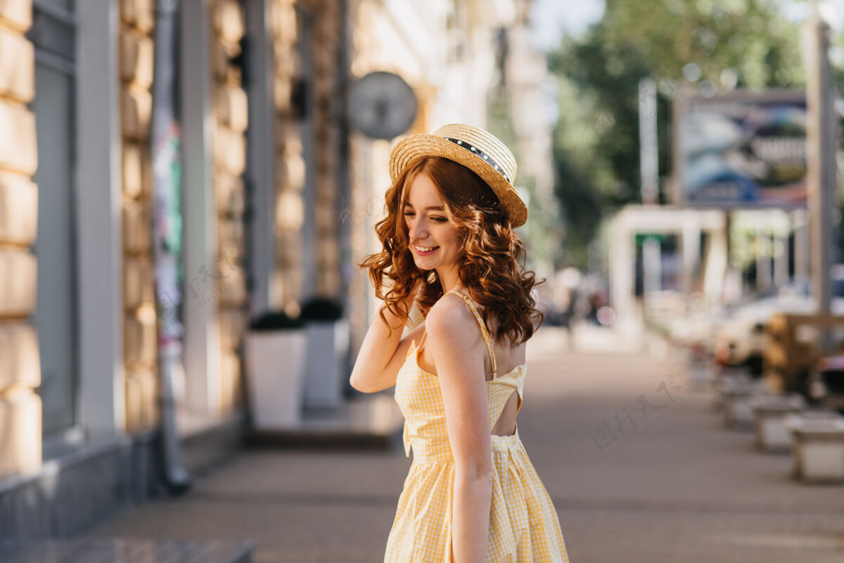 乐趣穿着黄色连衣裙的年轻女子在城市里愉快地摆着姿势戴着帽子的漂亮女孩在散步时欣赏照片的户外照片卷发波浪发成人