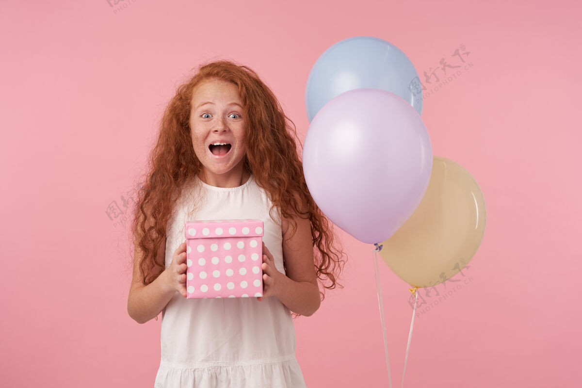 肖像照片中快乐的小女孩 红色卷发 穿着优雅的白色连衣裙 在粉色背景下摆姿势 拿着礼盒 为生日礼物感到兴奋 表达了真正的积极情绪卷曲欢快头发