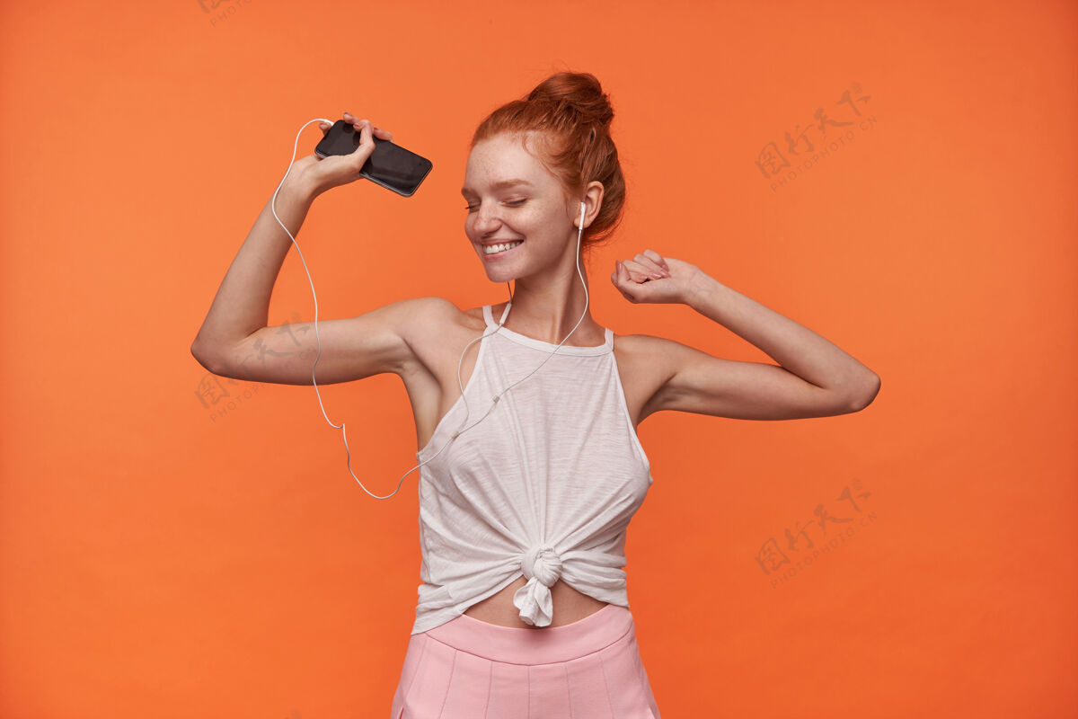 自然迷人的年轻女子戴着红色的头发在橙色的背景下打着结 闭着眼睛欣赏音乐曲目 举起手跳舞的摄影棚照片欢快积极室内