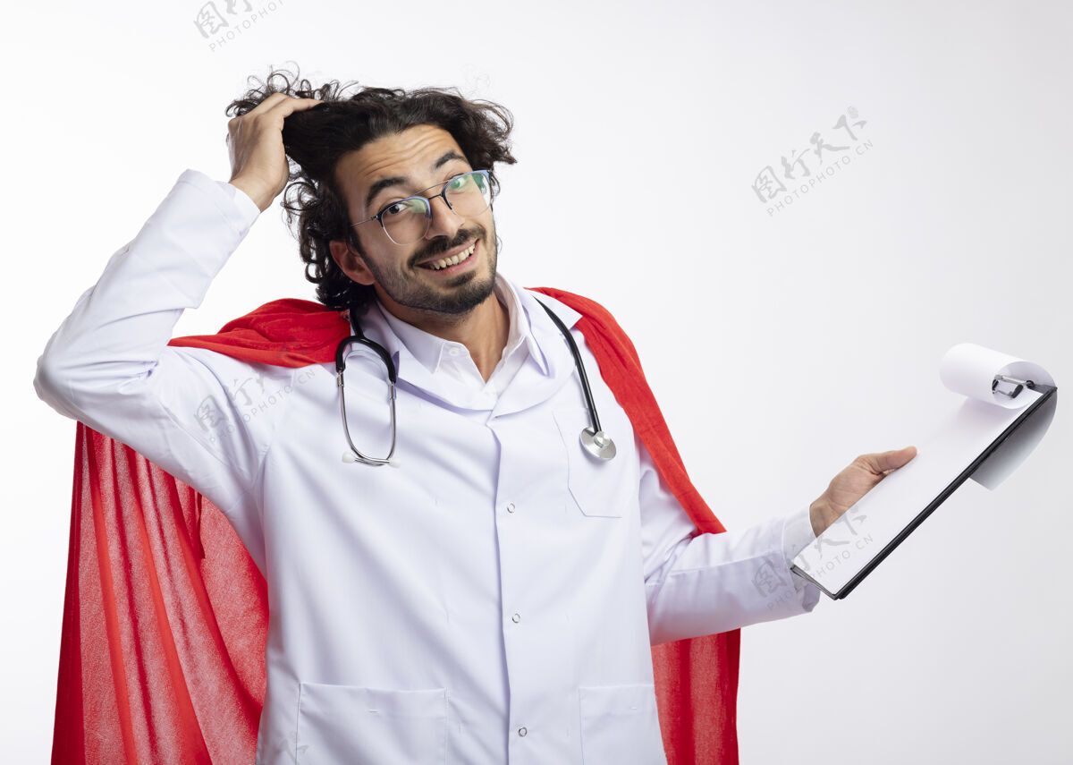 手戴着眼镜 身穿医生制服 披着红色斗篷 脖子上戴着听诊器 面带微笑的白人年轻人用手撩起头发 把剪贴板放在白墙上电梯持有医生