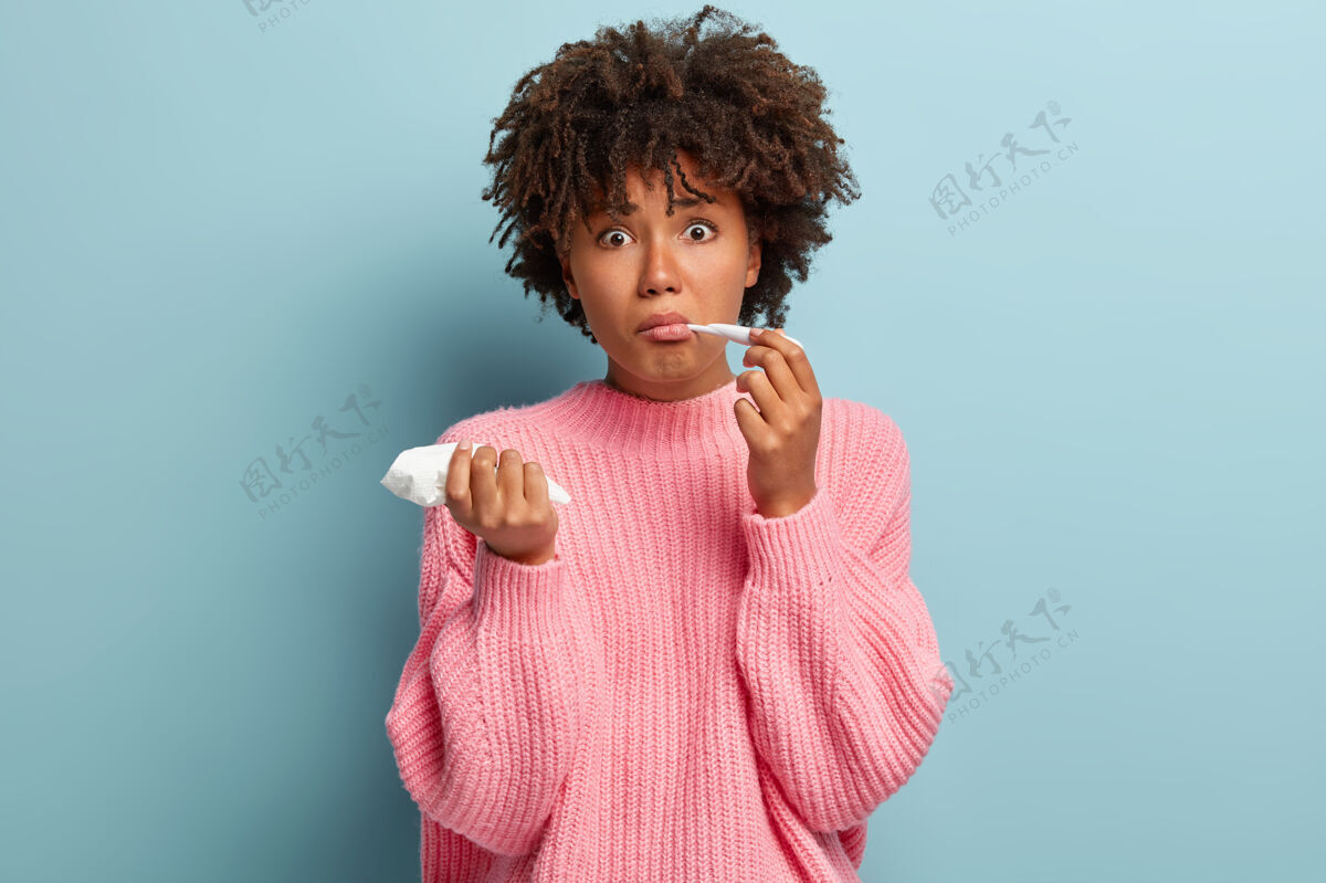 头部担心美丽不健康的女人感冒或发烧 测量体温 拿着纸巾 对流感感到惊讶 穿着超大的粉色套头衫 把温度计放在嘴里 隔离在蓝色的墙上组织跳楼成人