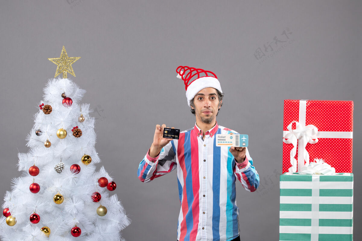 年轻前视图年轻人拿着卡片和旅行票绕着圣诞树和礼物男性肖像人