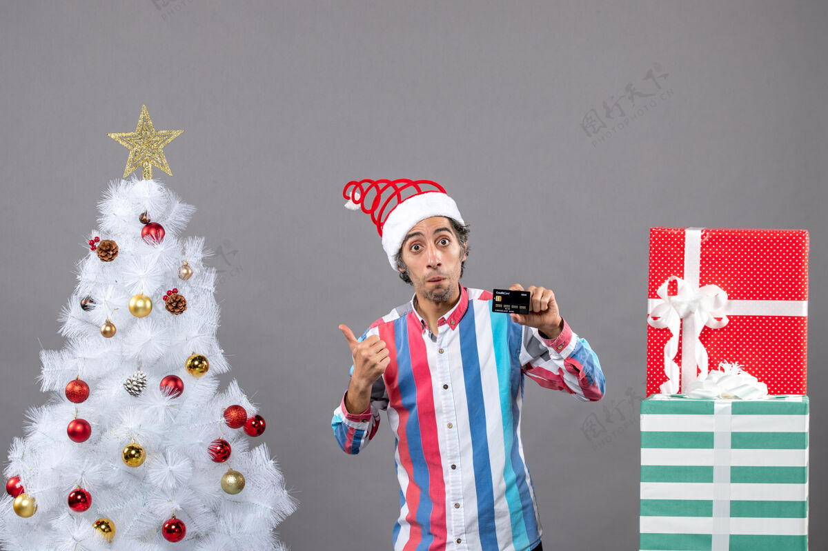 无辜前视图惊讶的男子信用卡显示圣诞树站在不同的礼物附近火炬礼物人