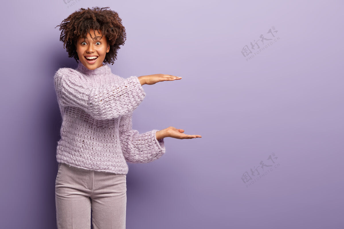 高兴高兴的非裔美国妇女的镜头做大小手势 演示了一些大 有卷发 穿着针织套头衫和裤子 孤立在紫色的墙壁上模拟复制空间数量高兴表情