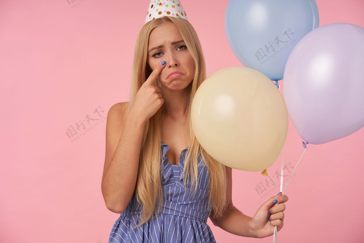 年轻心烦意乱的长发金发女人 穿着蓝色夏装 戴着生日帽锥形帽子 手里拿着五颜六色的气球 看着镜头伤心地擦去眼泪 孤立在粉色背景下金发娱乐女性