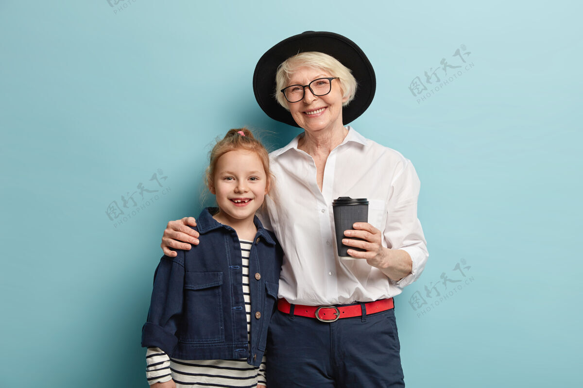 团结成熟满脸皱纹的奶奶和她学龄前的红发孙女紧紧地拥抱在一起 站在一起 微笑着 喝着外卖咖啡 有着良好的关系一代人的观念头饰一代女人