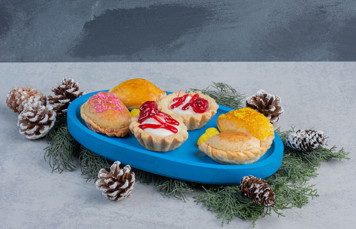 蛋卷蓝色盘子上的纸杯蛋糕 面包和果冻糖果 大理石表面装饰着松叶和圆锥体面包甜点果酱