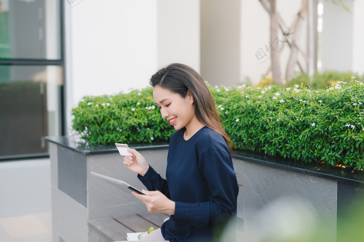 休闲一位年轻漂亮的亚洲女商人坐在办公楼外的长椅上咖啡信贷平板电脑