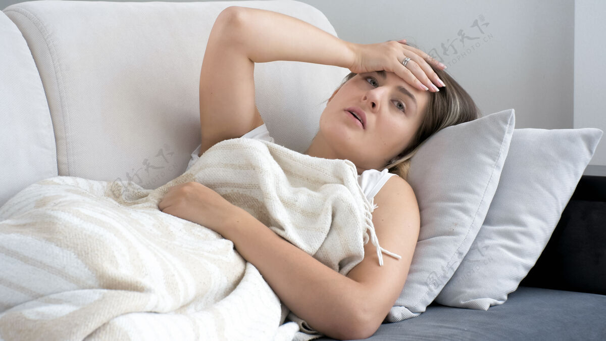 压力患有偏头痛的妇女躺在沙发上毯子下面的画像脸房子疲倦