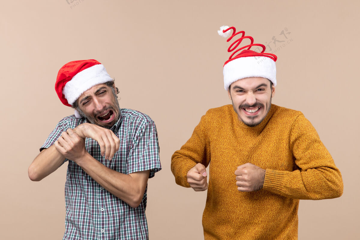 视图正面图两个戴着圣诞帽的家伙一个痛苦地握着他的手在米色孤立的背景上帽子痛苦米色