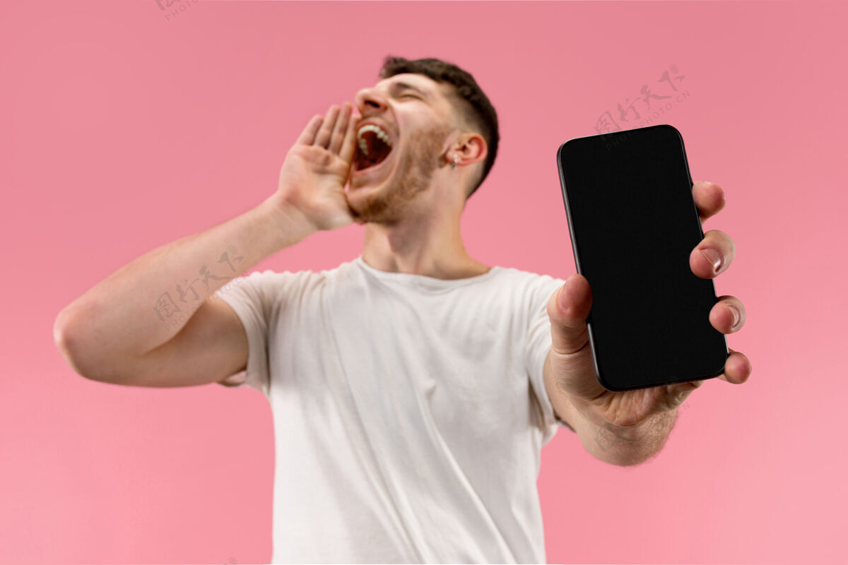 手机年轻帅哥在粉色背景下展示智能手机屏幕 惊喜万分屏幕表情指着