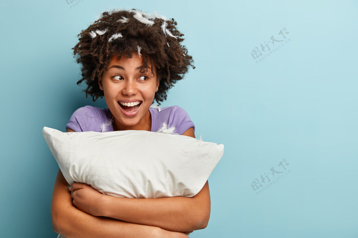 睡眠乐观的黑皮肤女人的照片 卷发 抱着柔软的枕头 午睡后心情很好 头上戴着羽毛 在蓝色的墙壁上摆姿势 空白处获取信息快乐风格高兴