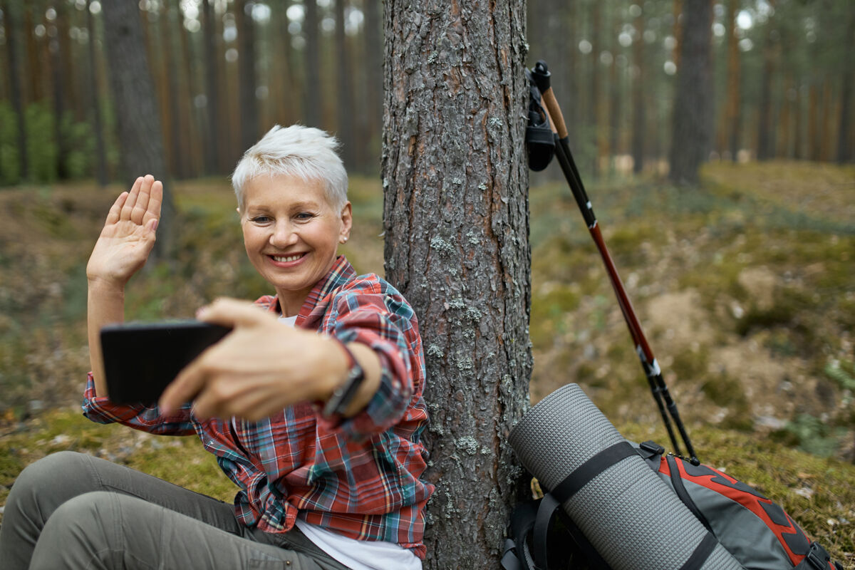 享受快乐活跃的退休女性坐在树下 带着远足装备手持手机 微笑着挥手 通过在线应用程序与朋友通过视频会议通话自然科技远足