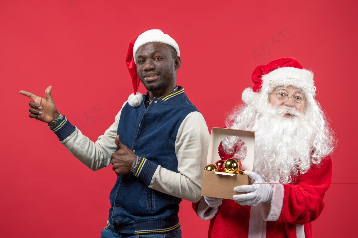 人圣诞老人和年轻人在红墙上拿圣诞树玩具的正视图圣诞快乐圣诞举行