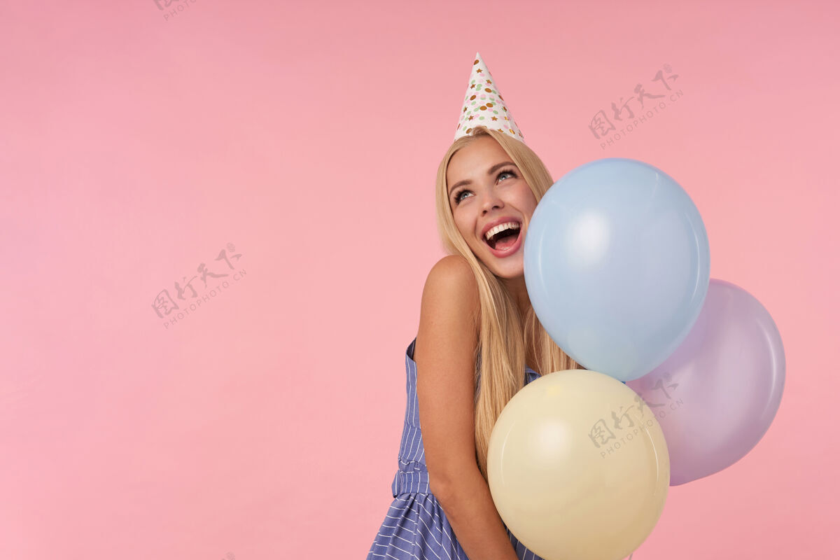 帽子美丽欢快的年轻长发女士 穿着蓝色夏装 戴着锥形帽子 在粉色背景上摆出五颜六色的氦气球 在收到令人敬畏的礼物时表现出愉快的反应女士空气五彩